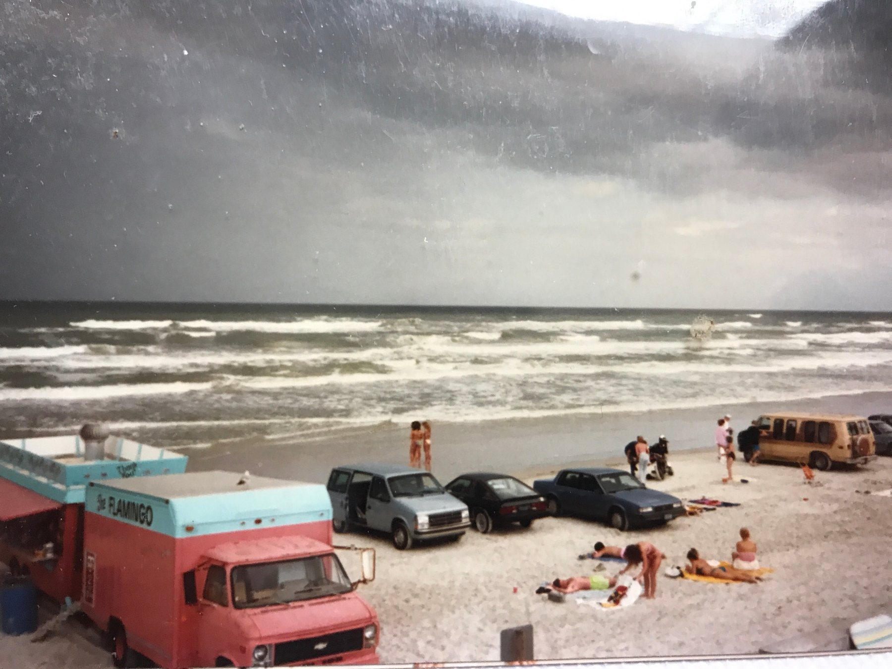 Beachgoers and vehicles beside rough surf under dark storm clouds